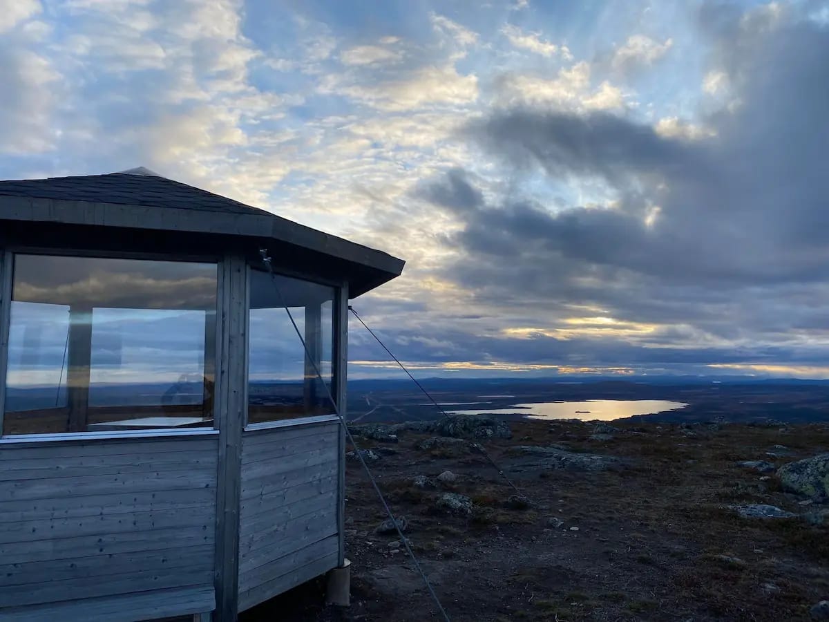 En fjällstuga på Dundret med reflekterande fönster fångar det dramatiska skymningsljuset över ett landskap med vatten och vidsträckta vidder.