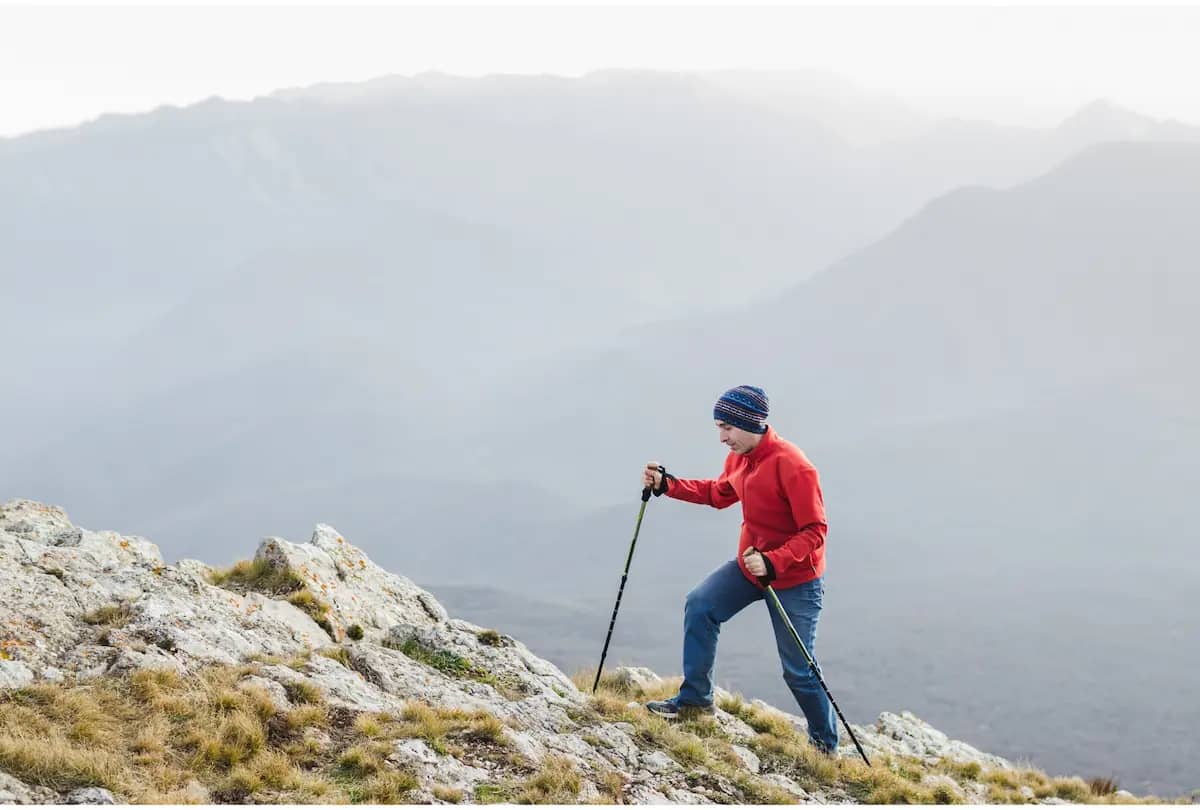 En ensam vandrare i röd jacka och randig mössa använder vandringsstavar på en klippig bergstopp med suddiga berg i bakgrunden.