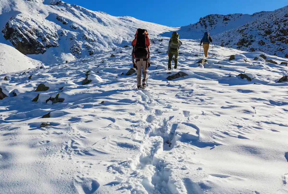 Grupp av tre vandrare går på en snöklädd bergsstig under en klarblå himmel.